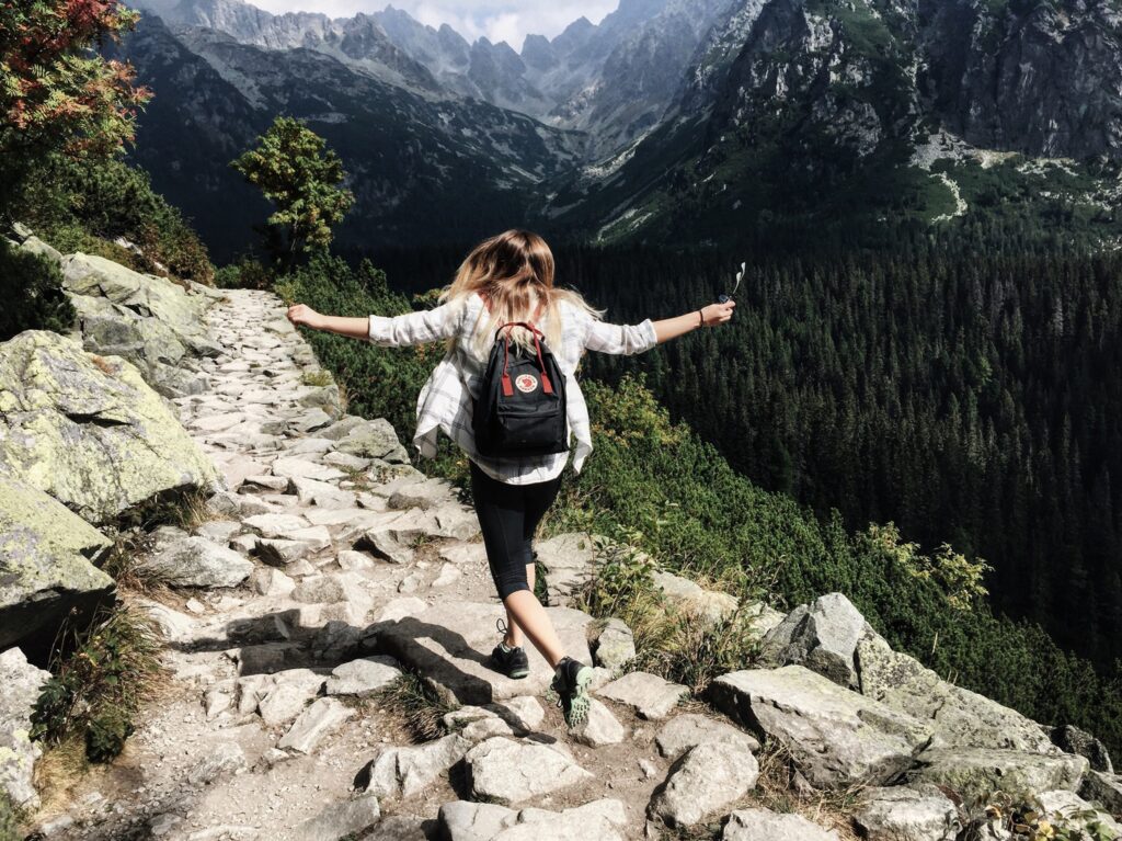 Woman wearing a backpack jumping along a rocky path through the mountains with trees filing the valley.